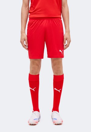 Bărbat purtând pantaloni scurți sport roșii Puma, șosete roșii asortate cu siglele Puma și ghete de fotbal albe pe un fundal simplu.