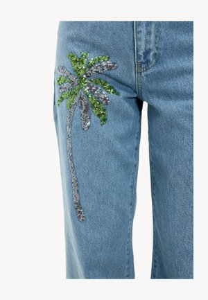 Un jean en denim bleu clair avec un design de palmier en sequins verts et argentés sur la jambe gauche, ajoutant texture et intérêt visuel.