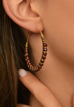 Des boucles d'oreilles en or en forme de créoles présentent des accents de perles rouges le long du bord, avec un fil texturé et une finition polie, mises en valeur sur une oreille.