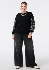 Zwarte sweatshirt met geborduurde bloemenpatronen op de mouwen, gecombineerd met wijde donkere jeans en lichtgekleurde studschoenen.