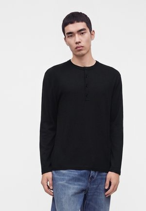Joven con camisa henley negra de manga larga y vaqueros azules, de pie sobre un fondo claro y liso.