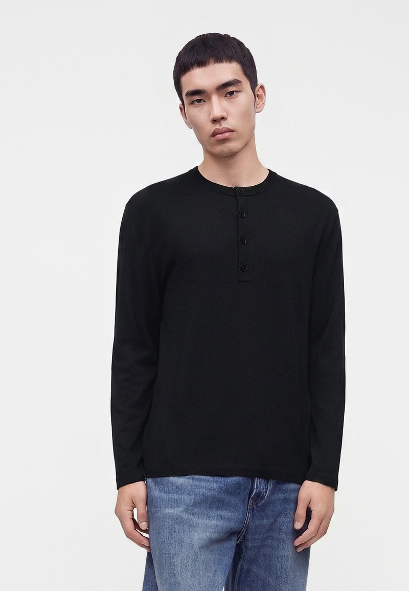 Jeune homme portant un t-shirt henley noir à manches longues et un jean bleu, debout devant un fond clair et uni.