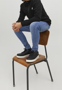 Zapatillas deportivas negras con suelas blancas, llevadas con unos jeans ajustados azules y una chaqueta negra con cremallera, sentado en una silla de madera.