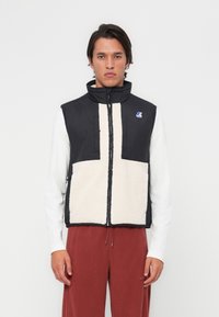 Gilet en polaire noir et crème avec un col haute, fermeture éclair sur le devant et logo sur la poitrine gauche. Présente des panneaux de tissu contrastants et un ourlet avec cordon de serrage.