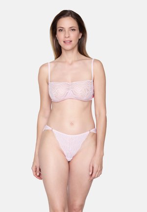 Vrouw staande, gekleed in een bijpassende lichtroze doorschijnende kanten beha en string, tegen een eenvoudige witte achtergrond.