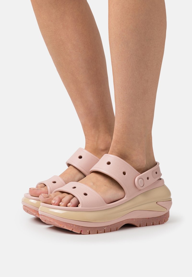 Pieds portant des sandales compensées épaisses roses avec des sangles réglables et des détails perforés, présentés sur un fond blanc.