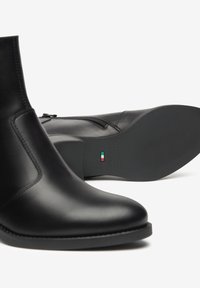 Een paar elegante zwarte leren enkellaarsjes, waarvan er één rechtop staat en de ander op zijn zijde ligt met de rubberen zool en het Italiaanse vlagdetail zichtbaar.