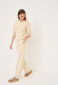 Felpa beige con cappuccio e logo arancione, abbinata a pantaloni della tuta beige coordinate. Il modello indossa sneakers marrone chiaro con dettagli grigi.