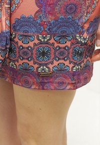 Gros plan sur un short à motifs colorés avec des dessins floraux bleus, violets et rouges porté sur une jambe, montrant une petite étiquette de marque "smash!".