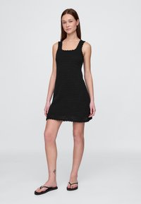 Crna kukičana haljina s kvadratnim izrezom, nazubljenim rubovima i opuštenim krojem. Model stoji na neutralnoj pozadini noseći crne japanke.