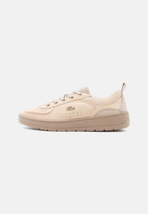 Beige suède sneaker met veters aan de voorkant, ademende perforaties, dikke zool en een trekkoord aan de hiel, weergegeven vanaf de zijkant tegen een witte achtergrond.