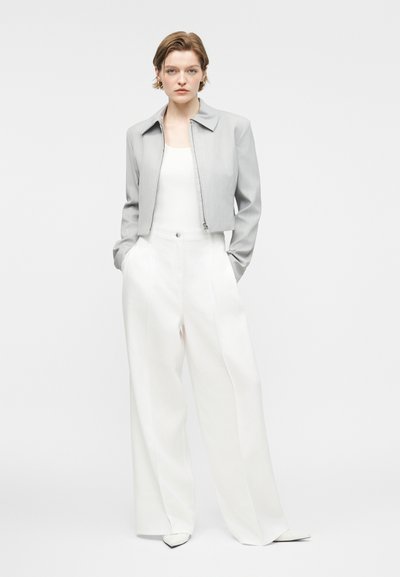Νέα γυναίκα με κοντά μαλλιά που φοράει ένα κοντό ανοιχτό γκρι σακάκι, λευκό τοπ, φαρδιά λευκά παντελόνια και λευκά μυτερά παπούτσια.