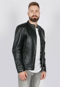 Chaqueta de cuero negra con cuello alzado, cierre frontal y dos bolsillos laterales. Llevada sobre una camisa blanca, combinada con jeans gris claro.