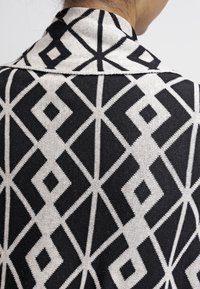 Tela de punto con un patrón geométrico en blanco y negro, con diamantes y líneas. El diseño incluye un cuello y una silueta estructurada.