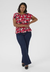 Top floreale rosso e nero con maniche corte, design a peplo e tessuto morbido, abbinato a jeans svasati blu scuro e scarpe nere.