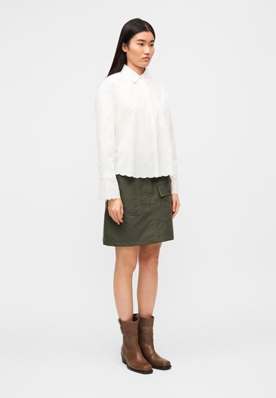 Λευκό πουκάμισο με κουμπιά, στεφάνη με κυματιστό τελείωμα και δαντελωτά μανίκια· σε συνδυασμό με φούστα ελιάς με τσέπες πατς· καφέ δερμάτινα μποτάκια αστραγάλου.