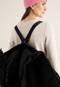 Zwarte buitenjas met een glanzende textuur, opgehangen met brede zwarte banden met de tekst "OUTERWEAR", gedragen over een lichtbeige trui.