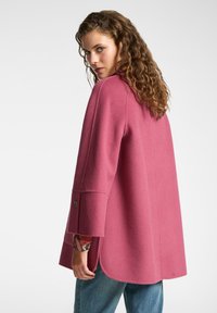 Cappotto in misto lana rosa con vestibilità rilassata, maniche ampie e dettagli con bottoni. Il retro presenta un orlo curvo e un tessuto testurizzato.