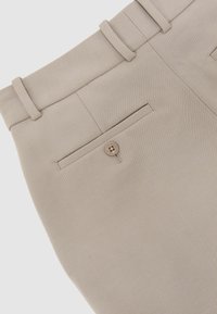 Bézs, szabott nadrág övbújtatókkal, hátul gombbal záródó nyílászsebből és látható varrási részletekkel.