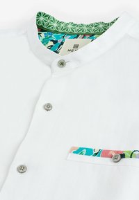 Chemise blanche avec un col mandarin, trois boutons gris, et des accents en tissu floral coloré sur la doublure du col et la bordure de la poche.