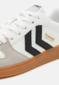 White leather sneaker with grey suede accents, black stripes, and a gum rubber sole. Features white laces and branding on the tongue.