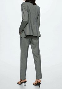Costume gris sur mesure avec une veste cintrée et un pantalon droit. Tissu léger avec une texture subtile, associé à des sandales à talons ouvertes.