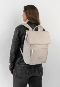Beige fabric backpack with a fold-over top, front zip pocket, and adjustable straps. Features a tag with the brand name "LARSON."
