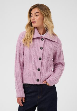 Femme aux cheveux blonds portant un pull rose en maille torsadée boutonné et un pantalon foncé, debout avec une main dans la poche, regardant sur le côté.