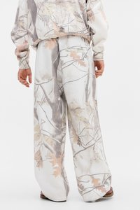 Pantalones deportivos blancos con un patrón de camuflaje que presenta hojas marrón claro y ramas grises; cintura elástica; tejido de algodón texturizado.