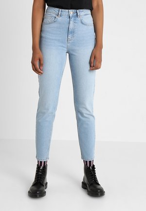 Person iført lyseblå højtaljede skinny jeans, stribede pink og sorte sokker, sorte snørebåndsstøvler og en sort top, stående på et ensfarvet gulv.