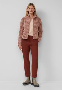 Vrouw in een roze textuur-ritsjas, lichtroze trui, roestkleurige broek en beige chunkybots tegen een effen muur.