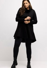 Tunic nera con cappuccio e orlo svasato, realizzata in tessuto morbido. Abbinata a jeans skinny neri e stivali neri chunky.