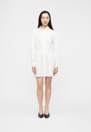Woman standing straight wearing a long-sleeve white shirt dress and black pointed shoes against a plain white background.