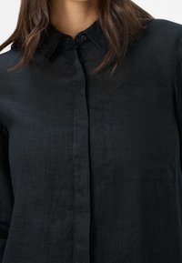 Chemise noire à manches longues avec boutons, en tissu texturé. Elle comporte un col et une poche poitrine.