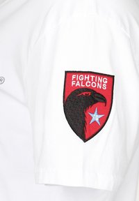 Witte t-shirt met een rood en zwart geborduurd patch met daarop een valk, de tekst "FIGHTING FALCONS" en een blauwe ster. Casual ontwerp.