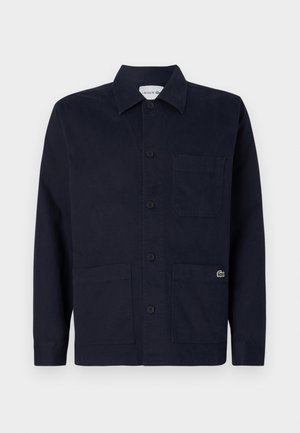 Chaqueta azul marino oscuro abotonada con cuello, tres bolsillos delanteros y pequeño logo verde de cocodrilo Lacoste en el bolsillo inferior derecho.