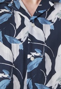Camicia a maniche corte con sfondo blu navy, caratterizzata da grandi modelli di foglie azzurro chiaro e bianchi e tre bottoni al colletto.