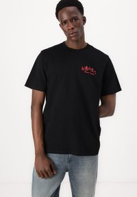 Zwarte katoenen t-shirt met rode grafische tekst op de borst, ronde hals, korte mouwen en een losse pasvorm.