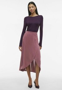 Un haut violet à manches longues associé à une jupe enveloppante rose avec une fente haute, fabriquée dans un matériau doux, accompagnée de talons aiguilles marron.