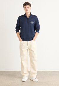 Felpa navy con zip e un piccolo logo, abbinata a pantaloni bianchi a vestibilità ampia e sneakers bianche, su uno sfondo neutro.