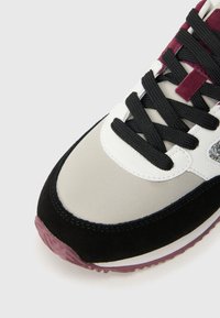 Primer plano de la puntera de una zapatilla con paneles en negro, blanco, beige y burdeos, cordones negros y una suela texturizada en color burdeos sobre un fondo blanco.