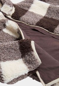 Chaqueta de forro polar de color marrón y crema con un cierre de cremallera, que presenta un exterior suave y texturizado y un forro interior de contraste.