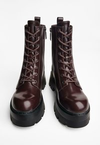 Botine din piele maro, cu șireturi, având o talpă robustă neagră, cu o textură netedă, ochiuri metalice și fermoare laterale pentru o îmbrăcare ușoară.