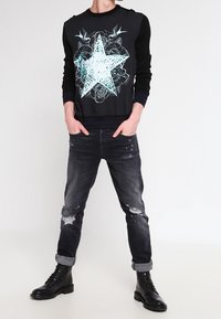 Black sweater with a large white star graphic, paired with distressed dark jeans and black lace-up boots. Casual fit, ribbed cuffs.