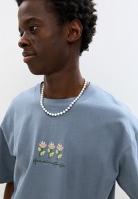 Blaues Baumwoll-T-Shirt mit floraler Stickerei in Pink und Grün, darunter der Schriftzug "Harmonie naturelle". Eine mit Perlen besetzte weiße Halskette sorgt für zusätzliche Details.