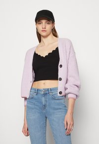 Cardigã canelado lilás claro com grandes botões pretos, usado sobre um top cropped preto com bicos, combinado com jeans de cintura alta azul claro.