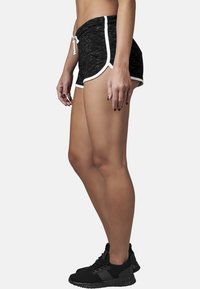 Černé sportovní šortky se strukturovaným vzhledem, s bílým lemem a šňůrkou na stažení. Nosené s černými teniskami v bočním profilu.