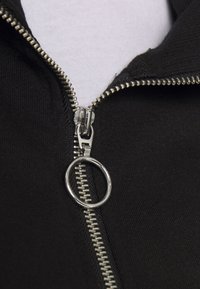 Giacchetto nero con zip e tirante rotondo argentato. Presenta una texture liscia e una cucitura visibile lungo la linea della zip.