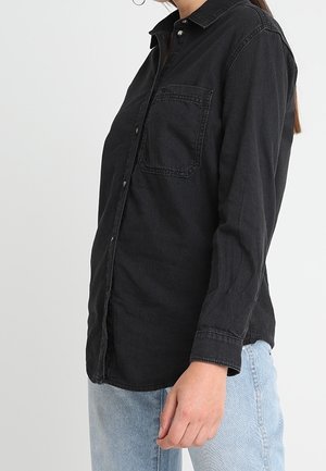 Vrouw draagt een zwart overhemd met lange mouwen, knopen en een borstzakje, en lichtblauwe spijkerbroek, staand tegen een effen achtergrond.