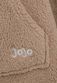Barna bolyhos anyag, amelynek puha, texturált felülete van. Jellemzője egy kis, fehér cérnával hímzett "Jojo" felirat. Zsebet tartalmaz.